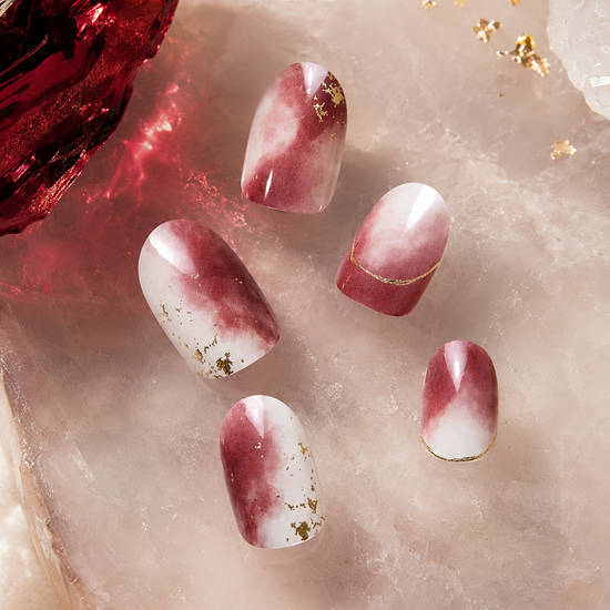 Five artificial nails with a white and deep burgundy marble pattern and small gold foil accents, displayed on a pink crystal slab next to a red crystal.